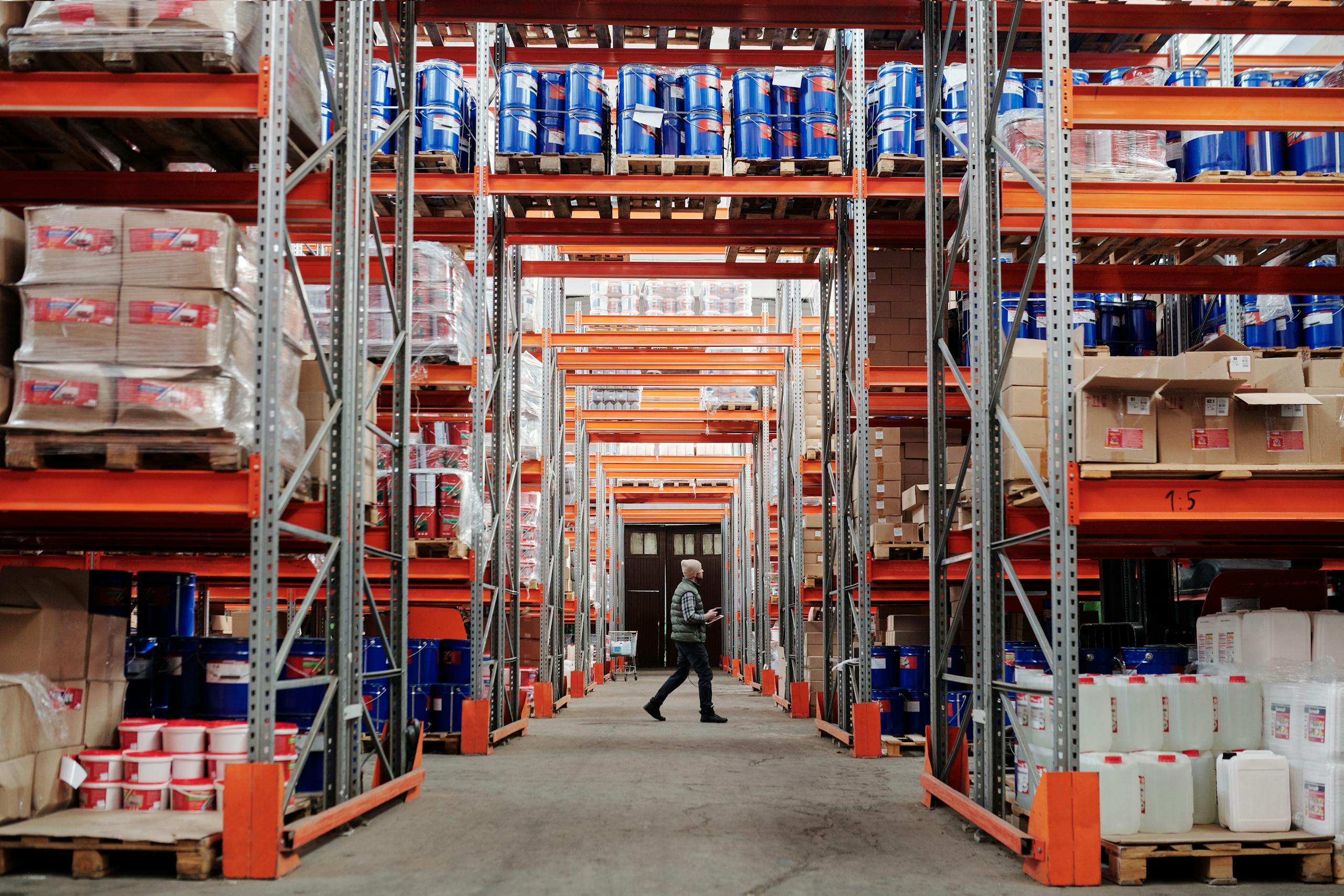 About Us A man walking through a large industrial warehouse with stacked shelves filled with goods and products.